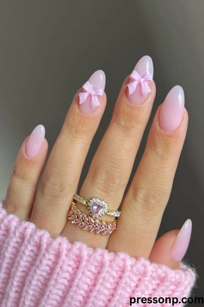 A close-up of a hand displaying mid-length, almond-shaped nails with a milky pink jelly finish, accented by 3D pastel pink bow charms on the middle and ring fingers.