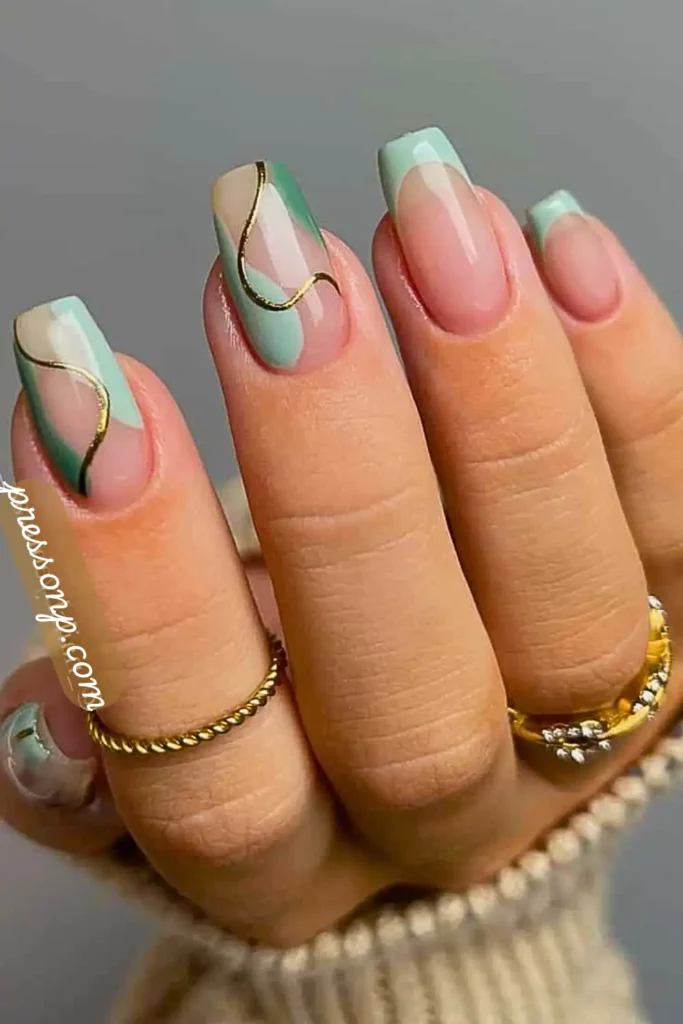 A close-up of a hand with long square nails showcasing a beach-inspired manicure with soft sage green French tips and abstract sea glass-like waves outlined in metallic gold foil.