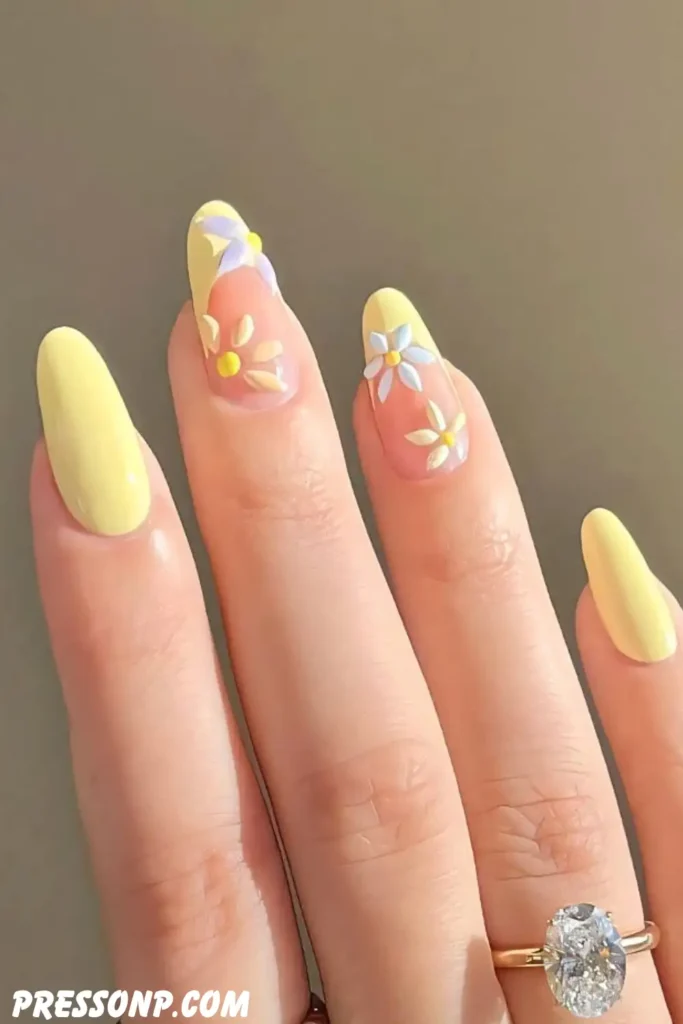 A close-up of a hand with elegant, long oval nails featuring a mix of solid soft butter yellow polish and clear nail bases decorated with delicate 3D white and yellow daisy petals.