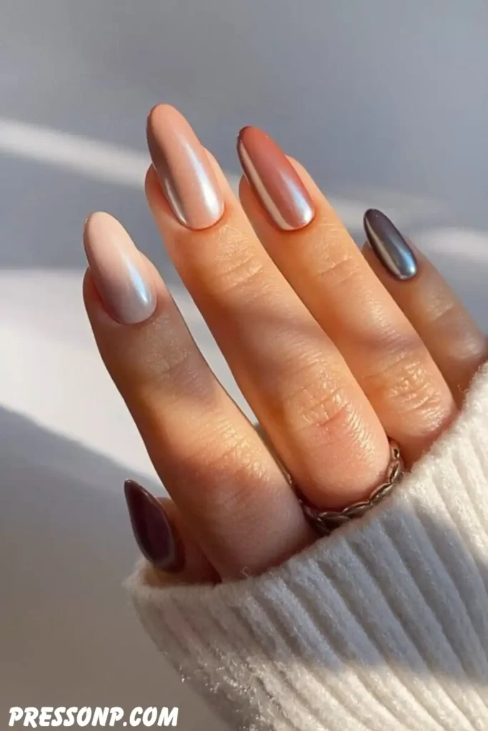 A close-up of a hand with long, elegant oval nails displaying a gradient skittle manicure of glazed chrome shades ranging from soft cream to warm brown.
