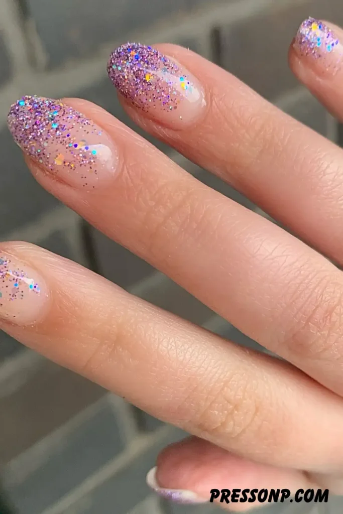 A close-up of a hand with long, elegant almond-shaped nails featuring a subtle glitter ombre design that fades from a sheer nude base to sparkling silver tips.