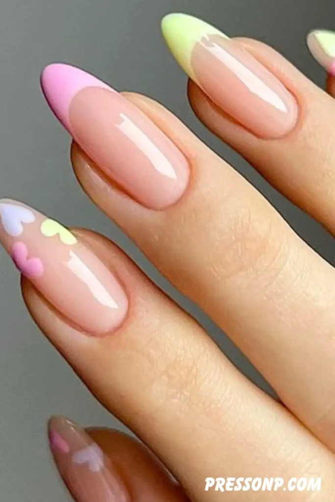 A close-up of a hand with long oval nails showcasing a modern pastel French manicure where each tip is a different soft shade, decorated with small pastel hearts on the accent nail.