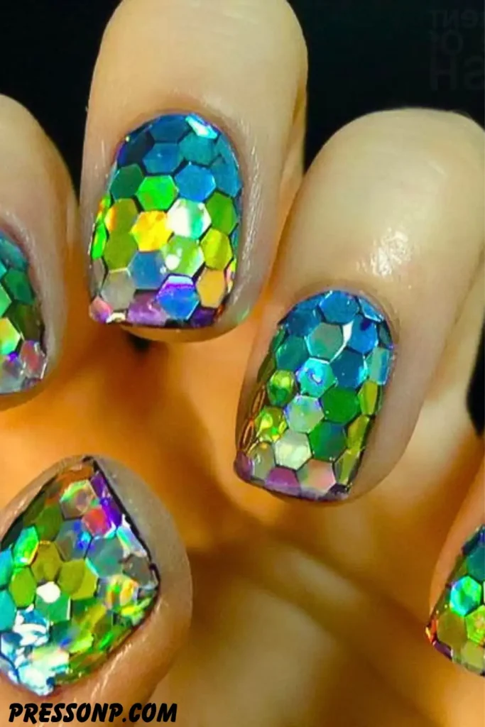A close-up of short, square nails completely covered in mosaic-style, oversized holographic hexagonal glitter that shifts through shades of green, blue, and gold to look like iridescent fish scales.