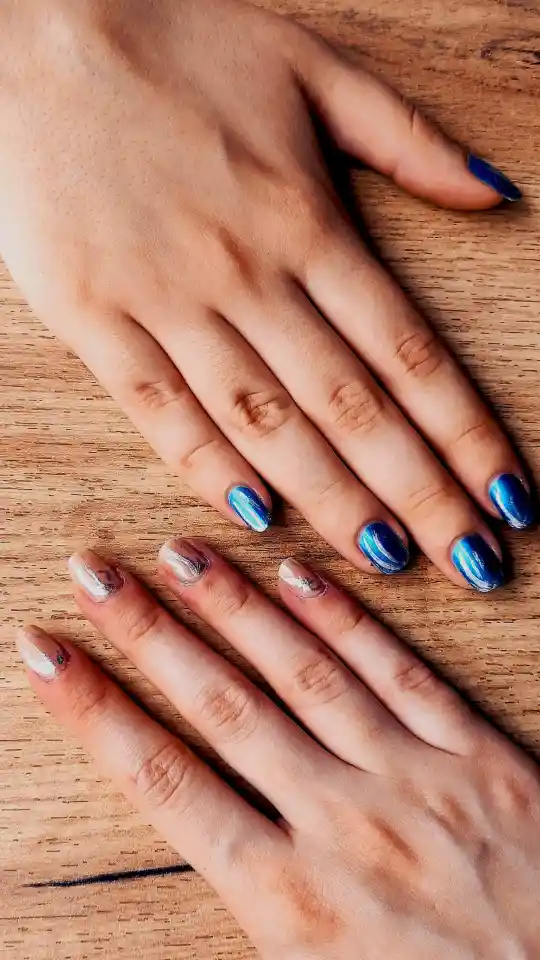 A top-down view of two hands resting on a wooden surface, showcasing a "cat eye" manicure. One hand features shimmering, multi-dimensional blue polish with a velvet-like finish, while the other shows a lighter, iridescent silver-blue design.