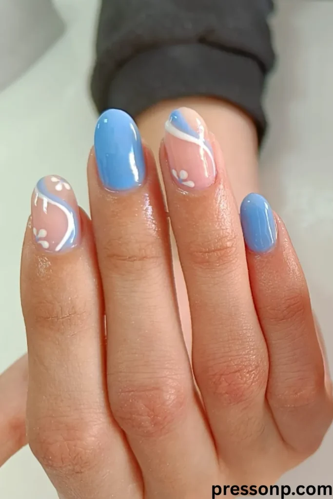 A close-up of a hand showcasing short, oval nails with a glossy sky-blue manicure combined with semi-translucent negative-space accent nails featuring fluid blue and white swirls and small white daisies.