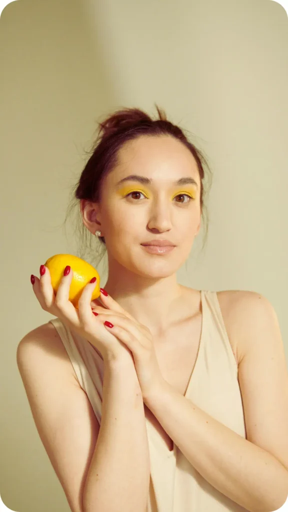A portrait of a woman with vibrant yellow eyeshadow, holding a bright yellow lemon. She has dark hair tied up, red nail polish, and wears a cream-colored sleeveless top against a neutral background.