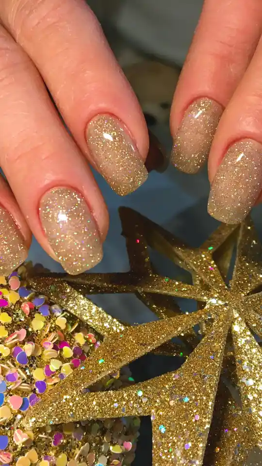 A close-up of fingers with square-shaped nails featuring a champagne-colored, glittery chrome finish. The hands are resting near a gold glitter star ornament and colorful heart-shaped confetti.