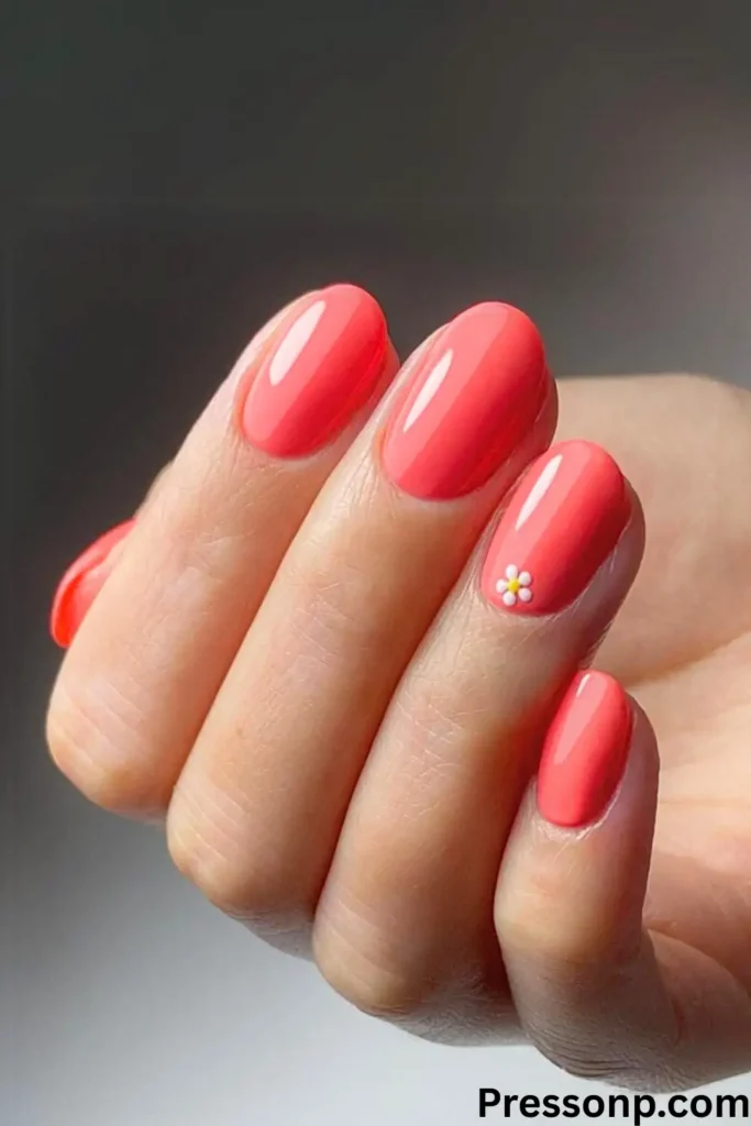 A close-up of a hand with vibrant coral-pink almond-shaped nails. A single, tiny white daisy with a yellow center is featured as a minimalist accent on the ring finger.Cute Nails for Vacation