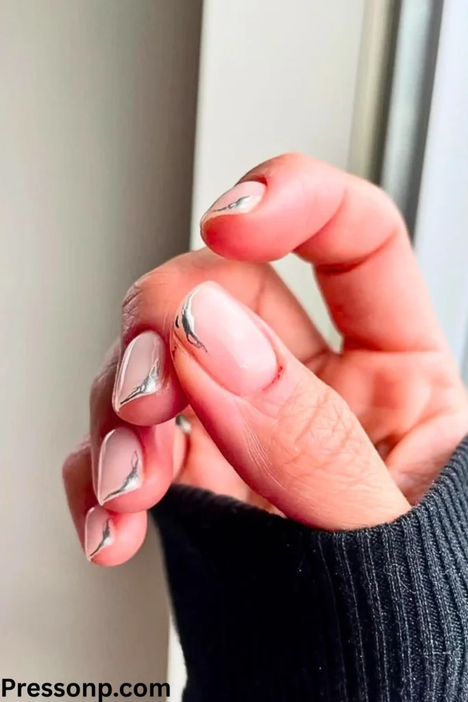 A close-up of a hand with almond-shaped nails featuring a modern micro-French manicure. The nails have a sheer nude base with ultra-thin, metallic silver tips that vary in shape on each finger.