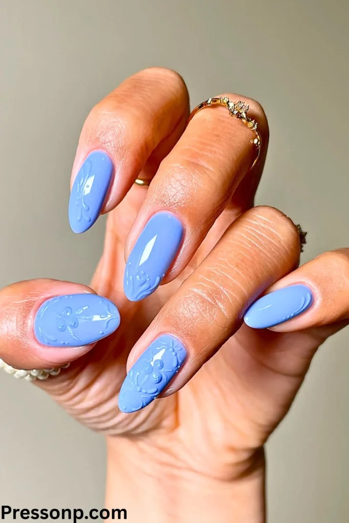 A hand with long, oval-shaped nails in a soft "blueberry milk" light blue. The nails feature intricate, 3D embossed floral patterns and are accessorized with delicate gold rings.