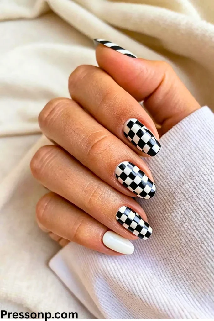 A hand featuring a bold checkerboard manicure. The almond-shaped nails showcase a classic black-and-white checkered pattern on three fingers, balanced by a solid white accent nail on the pinky.