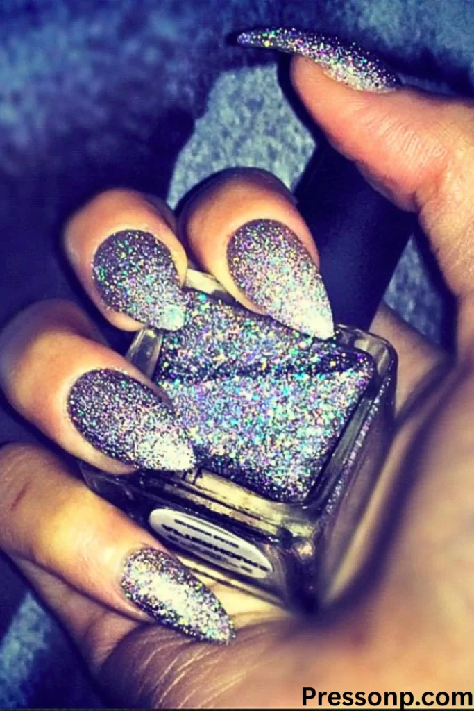A hand with long, stiletto-shaped nails completely covered in a dense, holographic silver glitter that reflects a multi-colored iridescent shimmer.