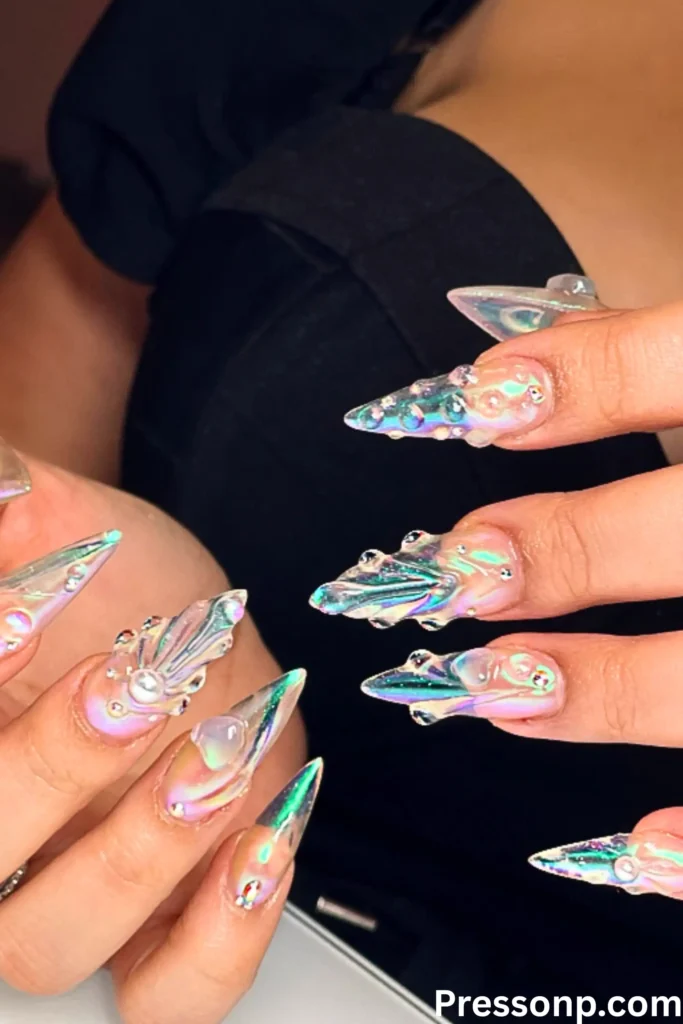 A hand with long stiletto-shaped nails featuring a translucent iridescent finish and 3D clear "water droplet" accents, creating a shimmering pearl effect.