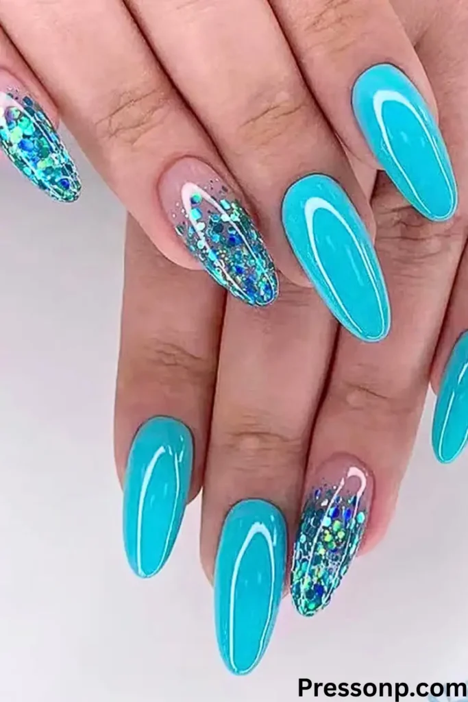 A close-up of hands with long, almond-shaped nails featuring a mix of solid bright turquoise polish and clear accent nails filled with sparkling blue and green chunky glitter.