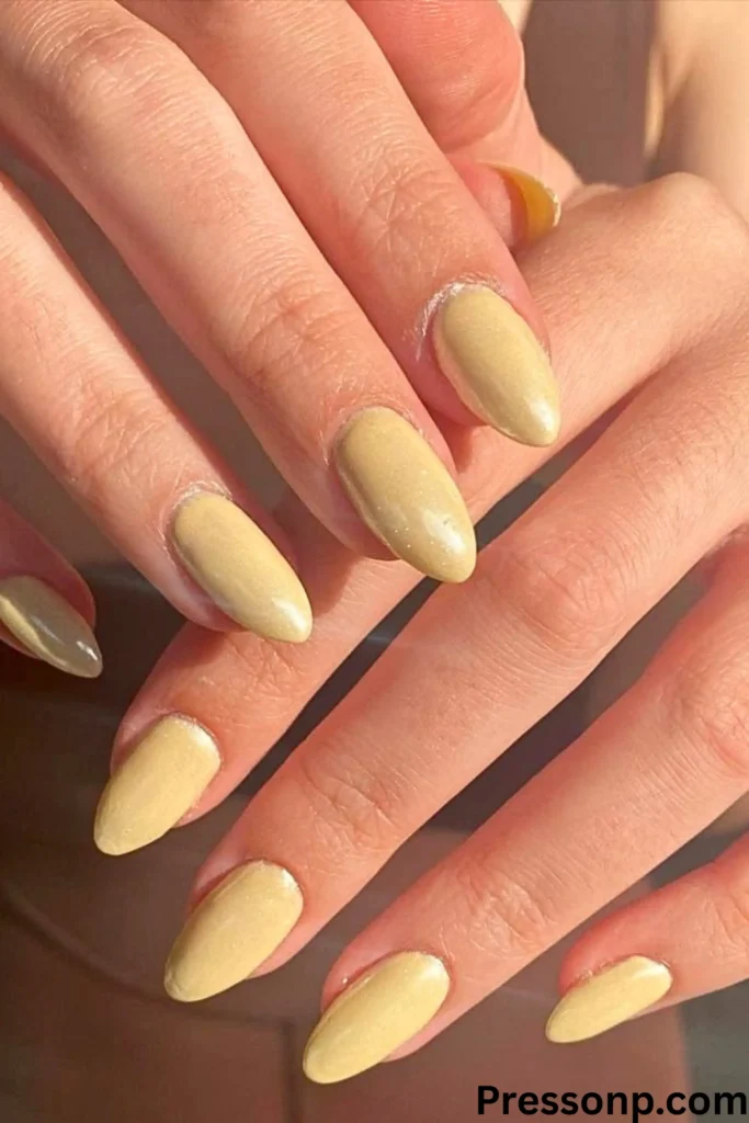 A close-up of two hands featuring a "Butter Yellow" manicure. The nails are a medium-length almond shape with a soft, creamy yellow polish that has a subtle pearlescent shimmer.