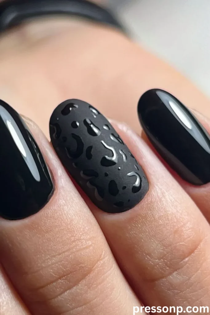 A close-up of long, oval-shaped black nails featuring an ultra-glossy finish, with a central accent nail styled in a velvety matte black base and detailed with raised, glossy black textures.