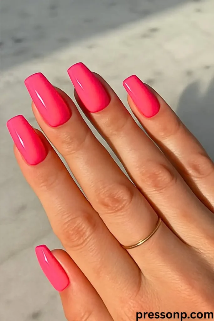 A close-up of a hand featuring long, glossy hot pink coffin-shaped nails and a simple gold midi ring, shown in bright natural light.