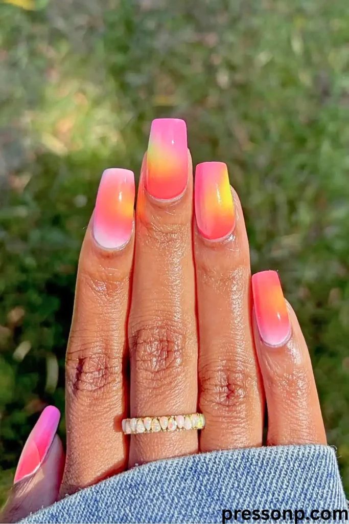 A close-up of a hand featuring medium-length square nails with a vertical neon ombré gradient blending bright yellow, orange, and hot pink, creating a sunset effect.