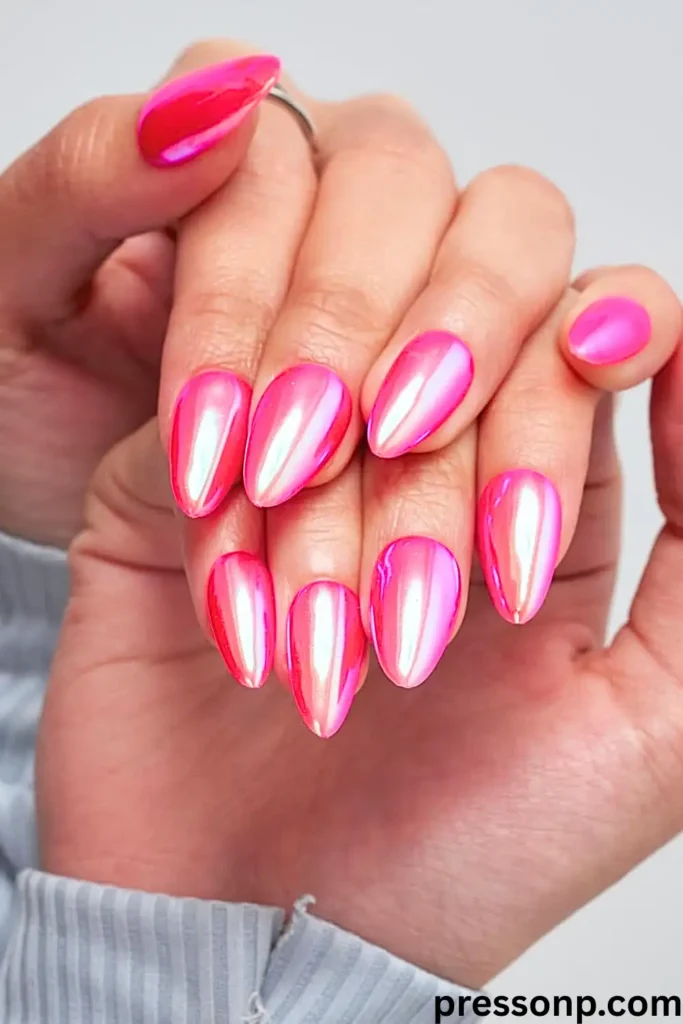 "A close-up of a person's hands showcasing a vibrant, futuristic manicure. The medium-length almond-shaped nails feature a fiery hot pink base finished with a high-shine iridescent chrome powder, creating a luminous, metallic glow that shifts from neon pink to pearlescent white in the light."