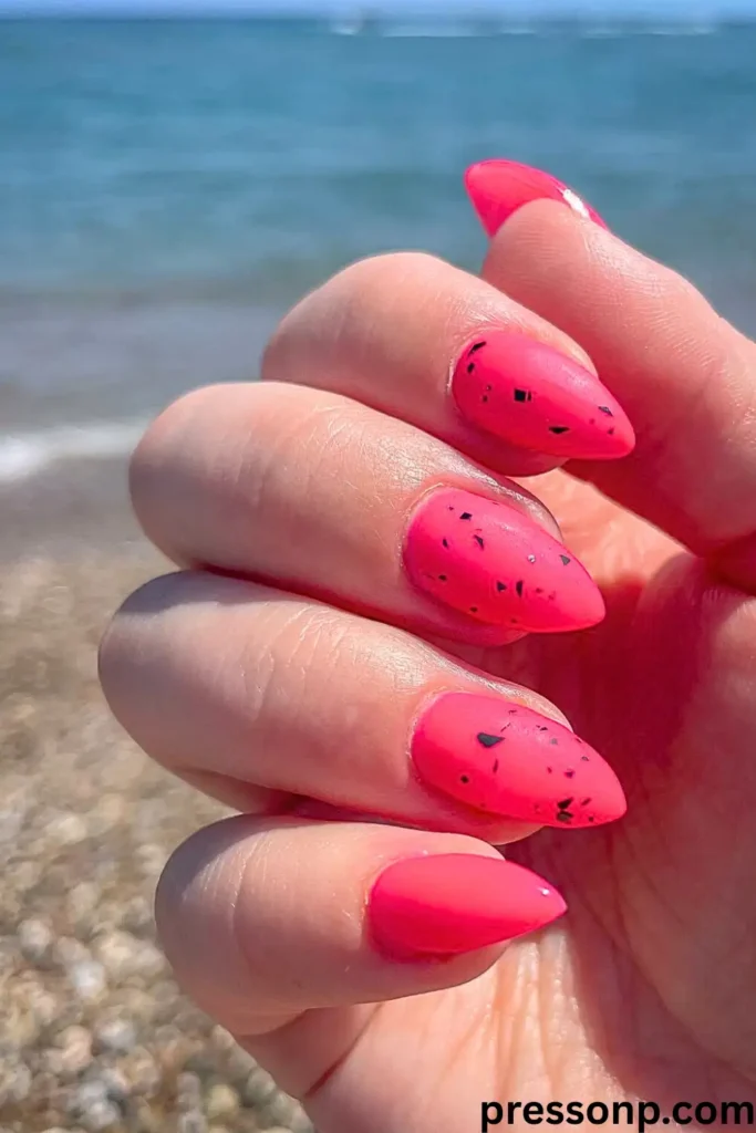 "A person's hand held against a sunny beach backdrop, showcasing almond-shaped nails in a vibrant matte hot pink. The middle fingers feature a modern art twist with small, black geometric flecks and lines scattered across the surface, resembling a stylish splatter or stone effect. The bold pink color and flat matte finish pop against the soft blue ocean and sandy shore in the background."