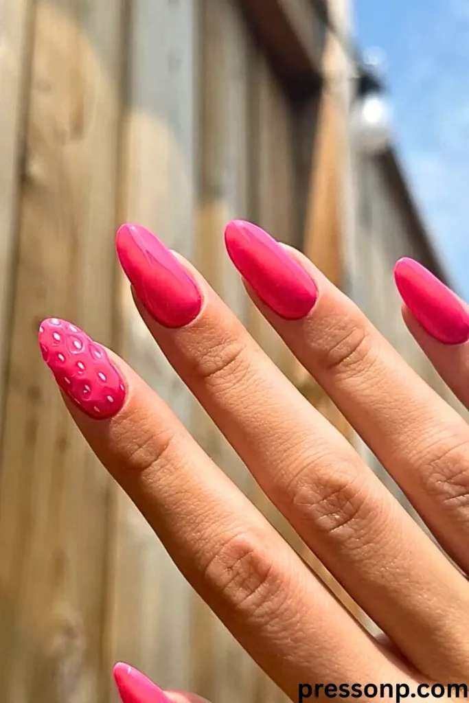 A close-up of a hand featuring vibrant, glossy hot pink almond-shaped nails, including a unique 3D strawberry-textured accent nail, shown outdoors in bright, natural sunlight.