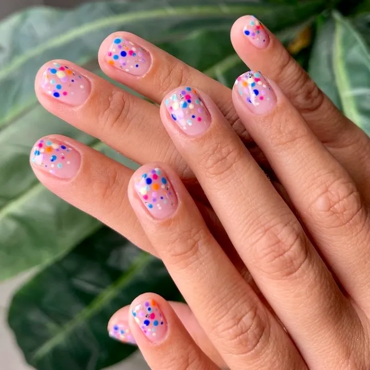 A close-up of short, square-oval nails with a glossy, transparent nude base coat decorated with scattered, colorful matte polka dots mimicking floating party confetti.