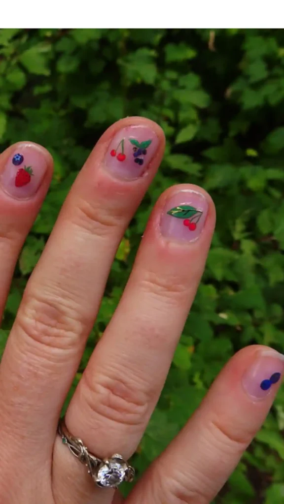 A close-up of a hand featuring "Micro Fruit" nail art. Each short nail is painted with a sheer pink base and tiny, detailed fruit illustrations, including strawberries, blueberries, and cherries, set against a background of green garden foliage.