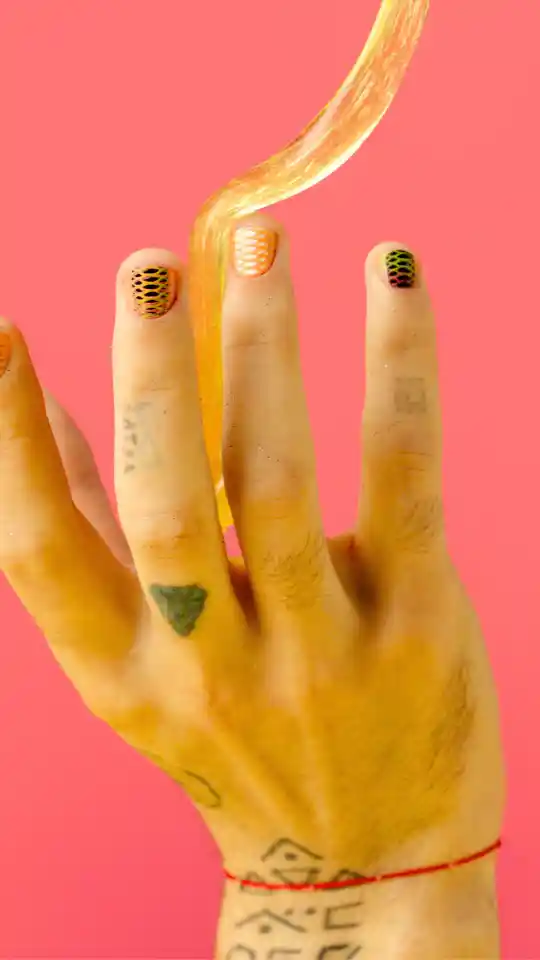 A hand with artistic patterned nail art held against a vibrant pink background. A thick, translucent golden liquid swirls between the fingers, complementing the warm yellow and orange tones of the manicure.