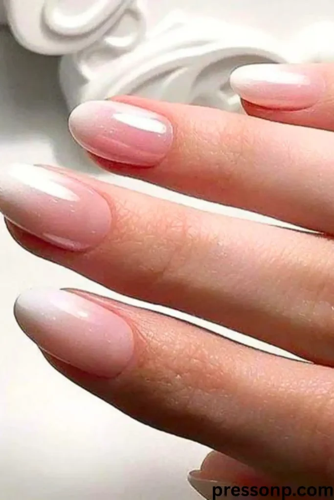 A close-up of a hand with long, elegant round-oval nails featuring a high-gloss, sheer nude-pink manicure with a soft, iridescent pearlescent shimmer.