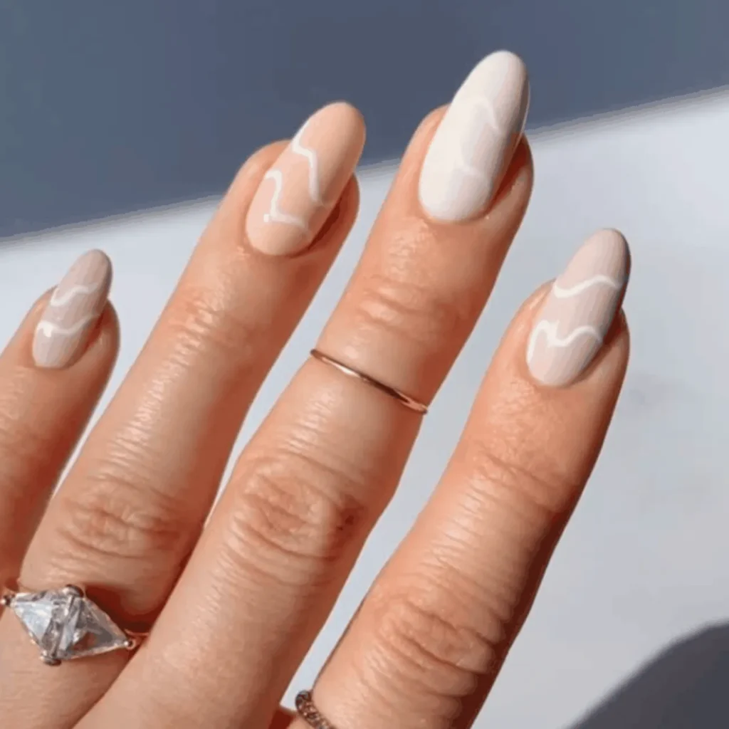 A close-up of long, oval nails painted in a mix of matte off-white and soft peach neutral tones, each decorated with a minimalist, fluid white wavy line flowing across the nail bed.