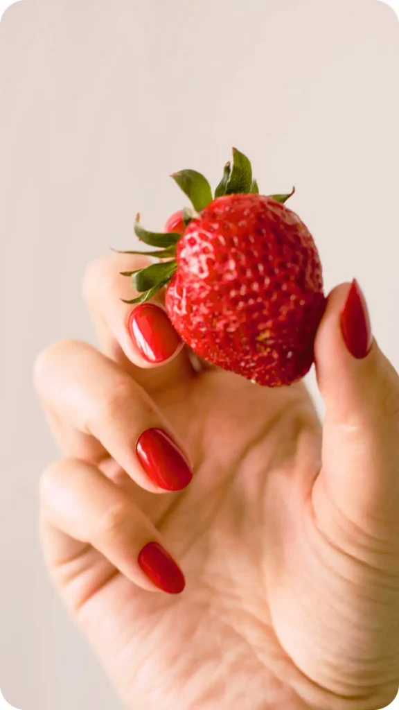 A close-up shot of a hand with glossy red manicured nails holding a fresh, ripe strawberry against a neutral, soft-focus background.
