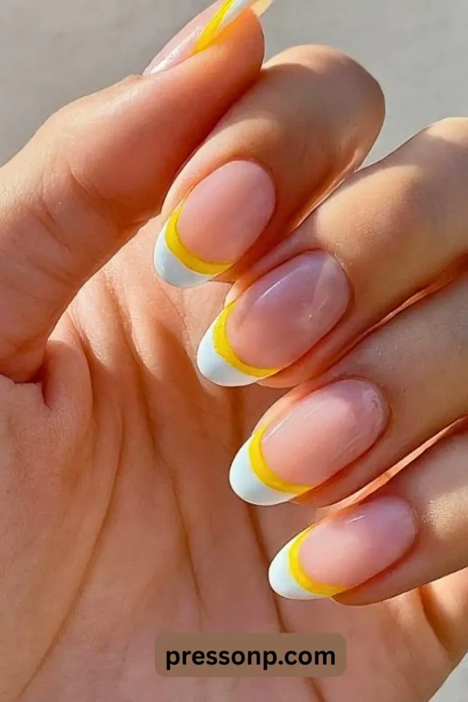 A close-up of "Sunshine French Tips – A Classic With a Twist" on elegant, almond-shaped nails. The design features a neutral nude base with a creative double-layered French tip: a bright sunshine yellow smile line topped with a crisp white edge. This modern take on a classic look offers a clean, cheerful, and sophisticated summer manicure.