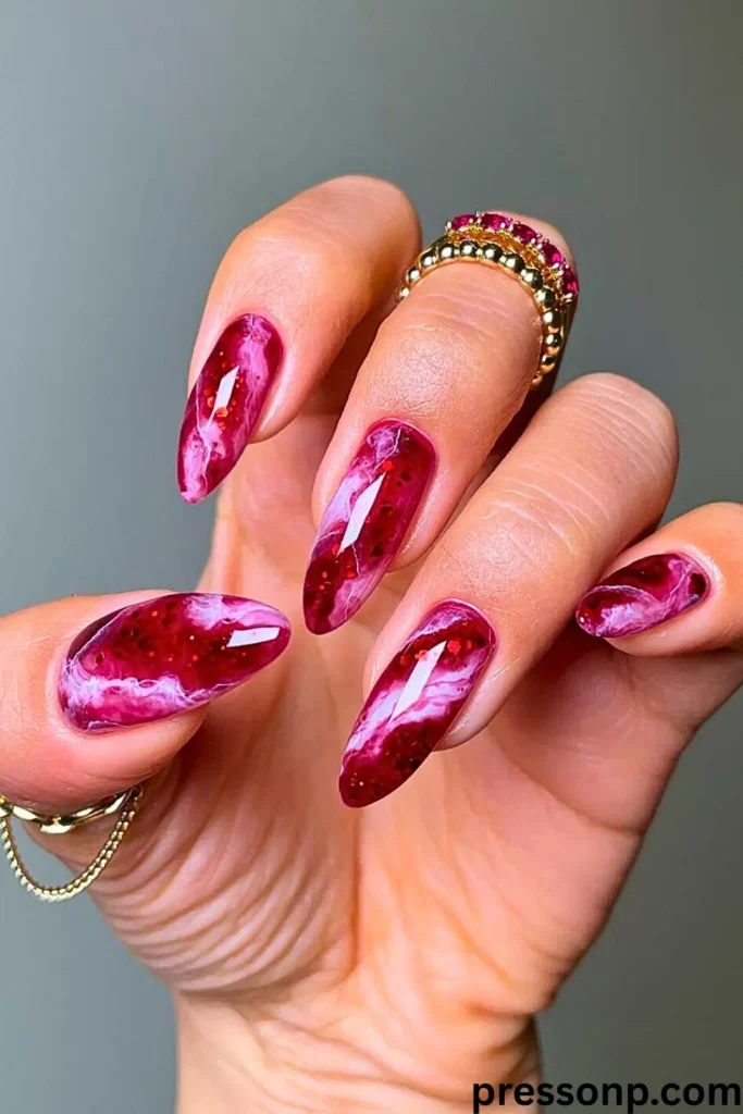 A hand showcasing "Sunshine & White Marble – Clean Meets Bold" nail art on long, almond-shaped nails. The design features a deep magenta and ruby red marble effect with wispy white smoke-like swirls and subtle glitter accents. The high-gloss finish is complemented by gold and ruby-encrusted rings, creating a sophisticated and bold summer look.