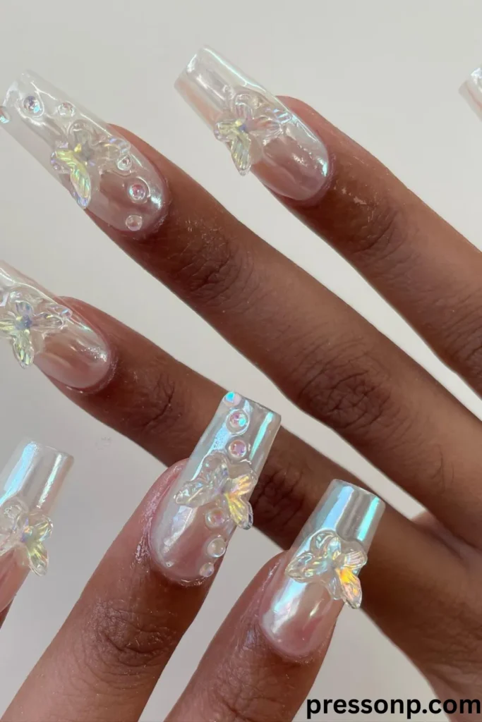 A close-up of a hand displaying long, square-shaped acrylic extensions with a luminous white iridescent chrome base, decorated with large 3D clear butterfly charms and matching clear rhinestone water droplet accents.