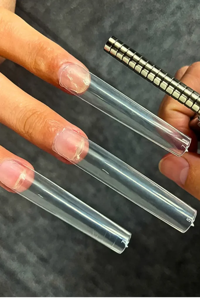 "A close-up demonstration of a time-saving nail hack: extra-long, clear square nail tips being applied to natural nails. A technician's hand holds a magnetic tool near the tips, illustrating a 'pro hack' for efficient extension application for Summer 2026."