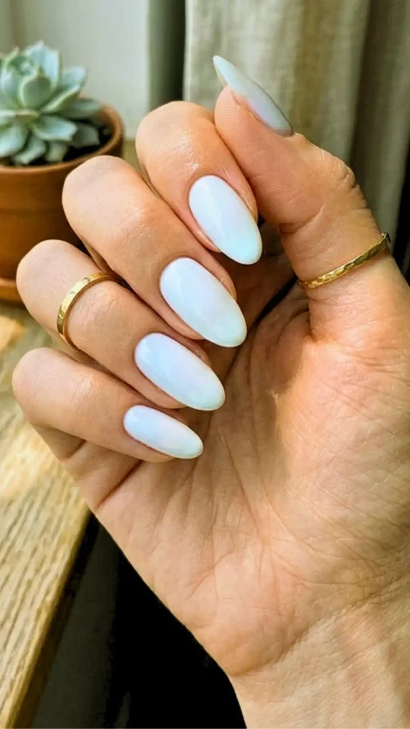 "A close-up of a hand featuring almond-shaped nails with a pearlescent white chrome finish, accessorized with thin gold bands, representing the 'Clean Girl' aesthetic for Summer 2026."
