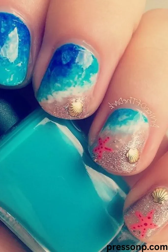 A close-up of short, rounded nails showcasing a detailed beach landscape manicure with ocean waves crashing onto sandy glitter beds, accented by tiny 3D gold seashells and pink starfishes.