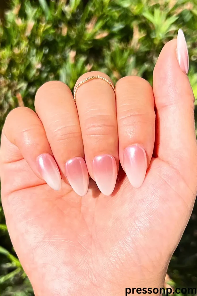 A close-up of a hand in bright sunlight showcasing long, elegant almond-shaped nails with a soft rose-pink to milky-white ombré gradient and a shimmering glazed finish.