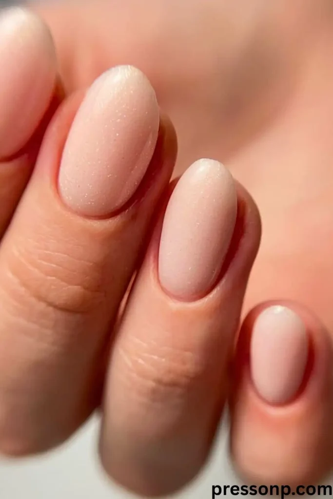 A close-up of short, neatly trimmed round nails painted in a simple, high-gloss solid pastel baby pink polish.