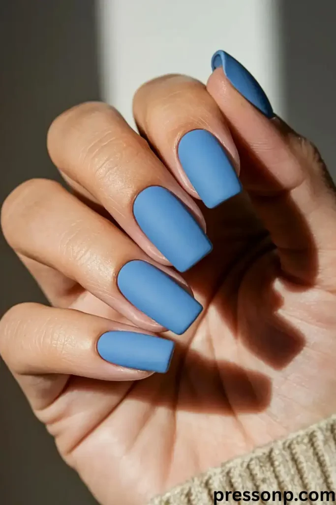 A close-up of a hand with long, elegant almond-shaped nails featuring a milky white base and abstract, fluid swirls in a soft dusty blue shade