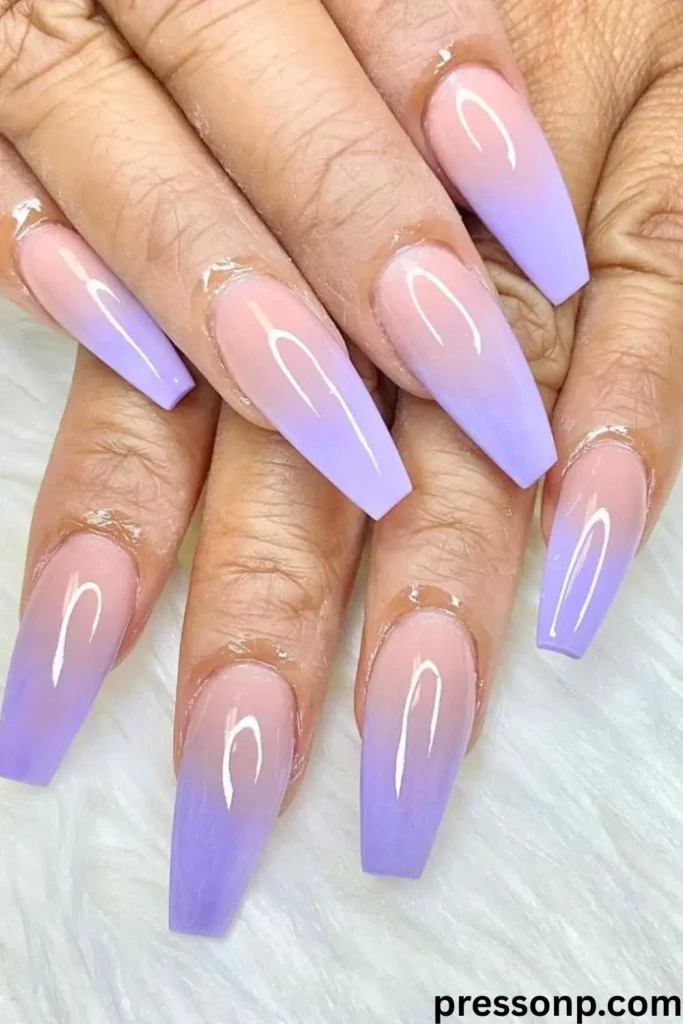 A close-up of hands displaying long, structured coffin-shaped nails featuring a smooth, vertical ombré gradient fading from a sheer nude base to a soft pastel lavender purple at the tips.