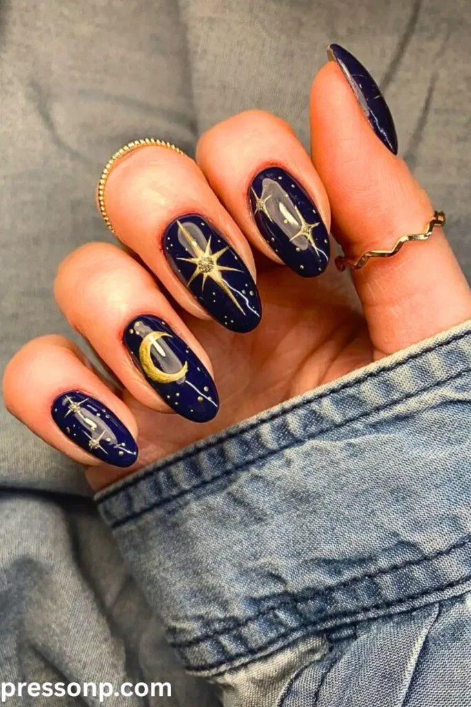 A close-up of long, oval-shaped nails featuring a glossy, deep navy blue base decorated with hand-painted gold crescent moons, starbursts, and tiny constellations.