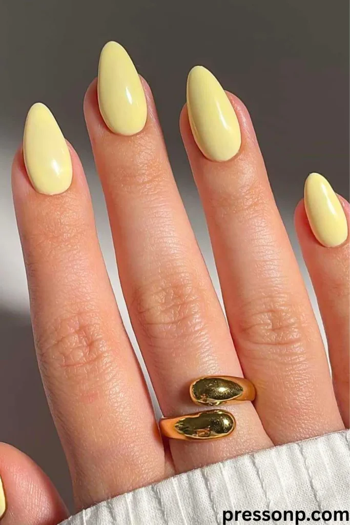 A close-up of a hand showcasing long, almond-shaped nails painted in a solid, glossy buttermilk pastel yellow color, accessorized with a chunky gold ring.