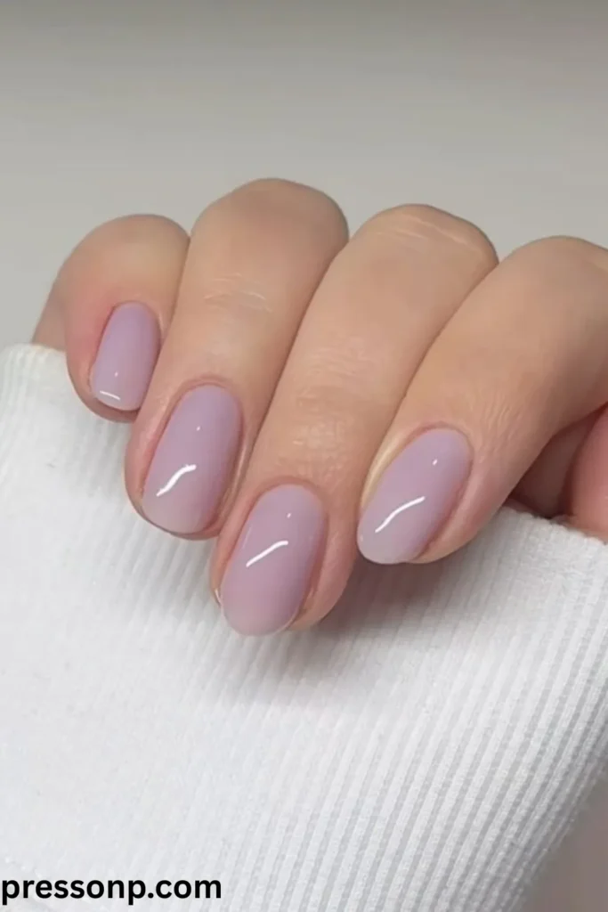A close-up of a hand showcasing a minimalist manicure on short, neatly filed squoval-shaped nails painted in a glassy, semi-translucent sheer lavender jelly purple polish.