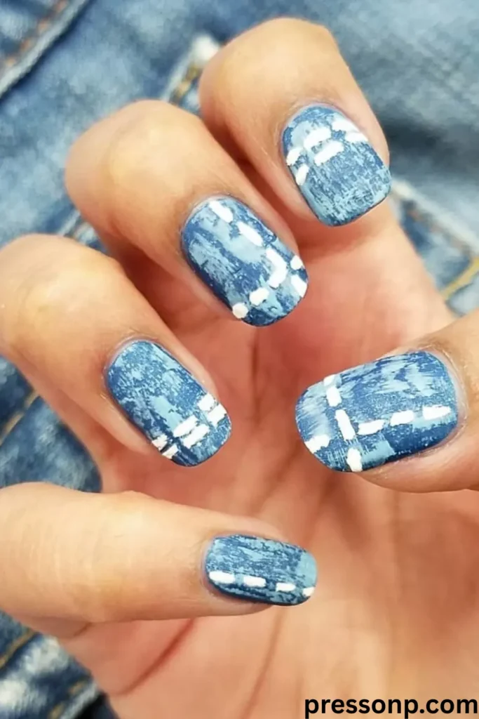 A close-up of a hand displaying short, square-shaped nails painted with a realistic blue washed denim texture, complete with hand-painted white stitching accents.