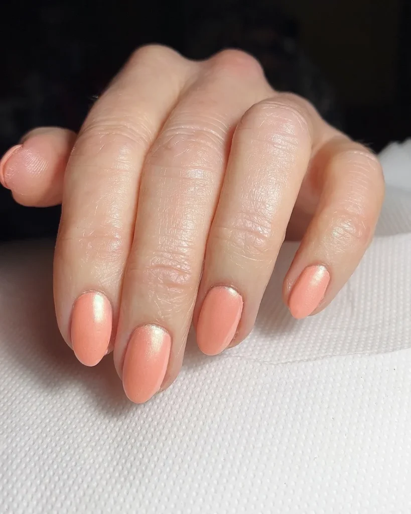 A close-up of a manicured hand resting on a white surface, showcasing short, neatly shaped oval nails painted in a soft peach color with a subtle satin-matte shimmer.