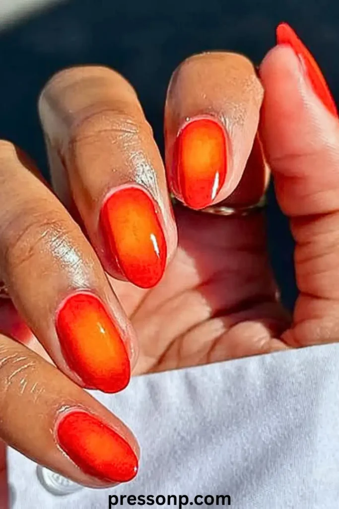 A close-up of a hand featuring vibrant red-orange almond-shaped nails with a glossy finish, styled against a crisp white shirt.