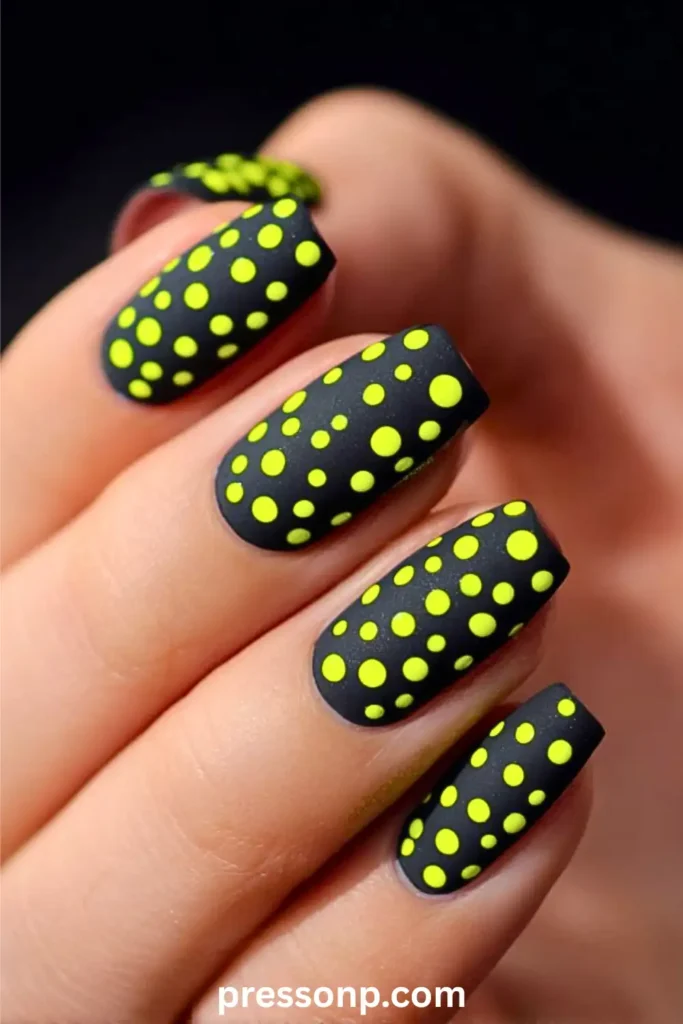 A close-up of a hand with short, square nails featuring a matte black base covered in various-sized neon yellow polka dots.