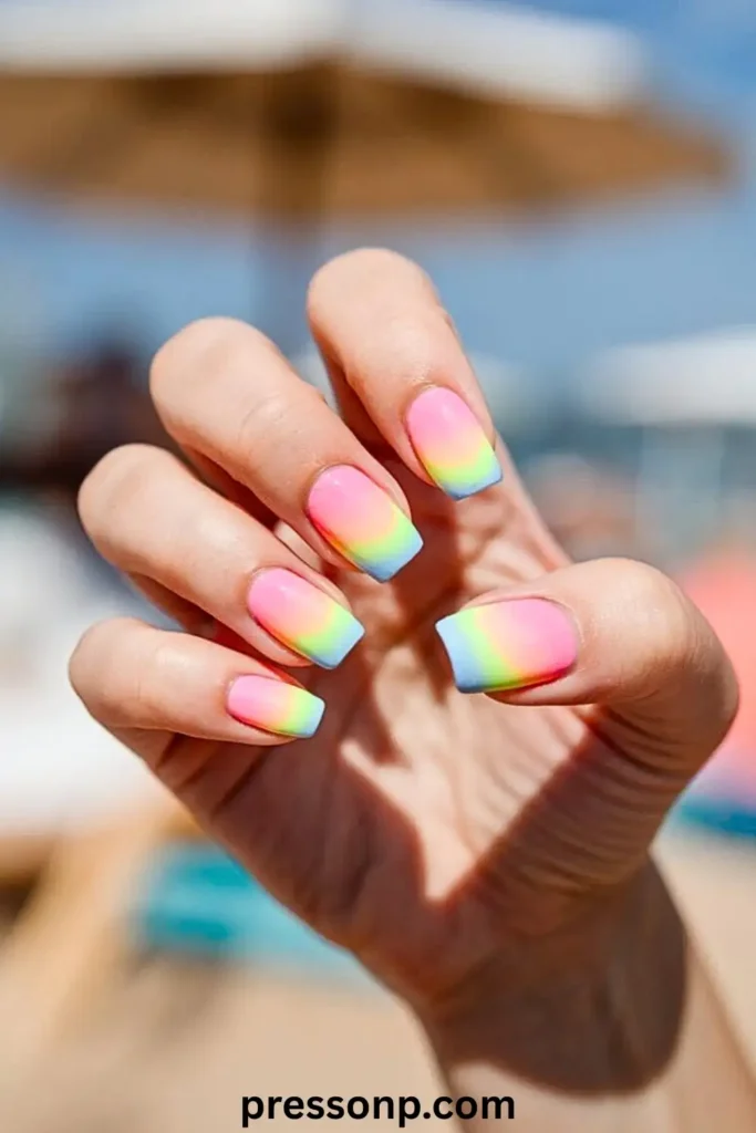 A hand showcasing medium-length square nails with a vibrant rainbow ombré French tip design, fading from pink to yellow, green, and blue, set against a sunny outdoor beach background.