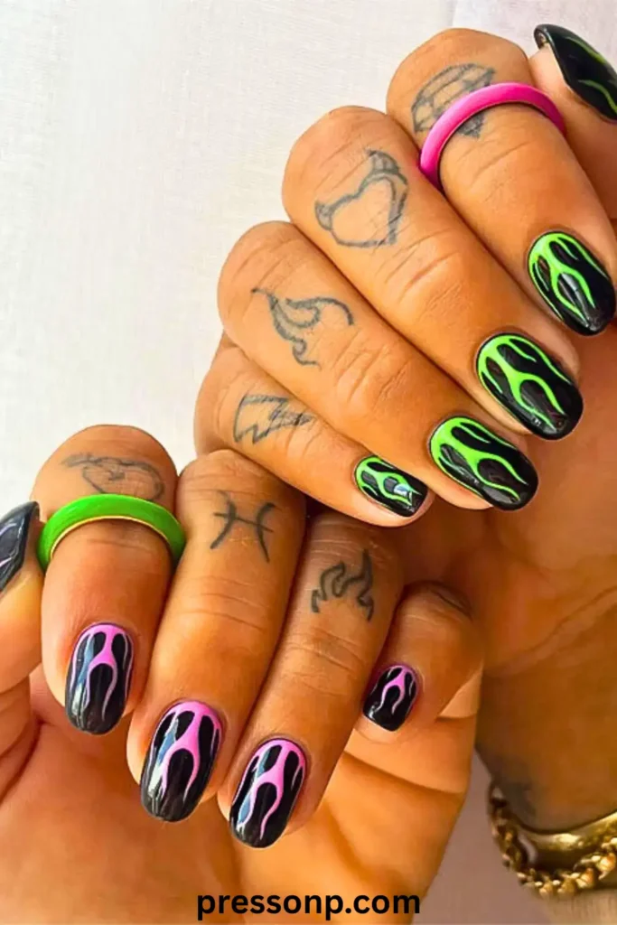 A close-up of two hands featuring a mix-and-match flame manicure. One hand has neon green flames on a black base, while the other features neon pink flames, complemented by small finger tattoos and matching colorful rings.